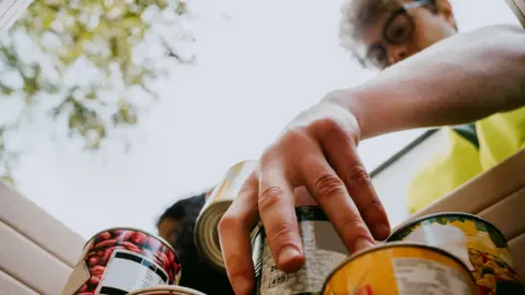 Getty Images A man wearing glasses is looking down at a box filled with canned food. He is wearing a bright yellow jacket and holding one of the cans. Trees are above him.