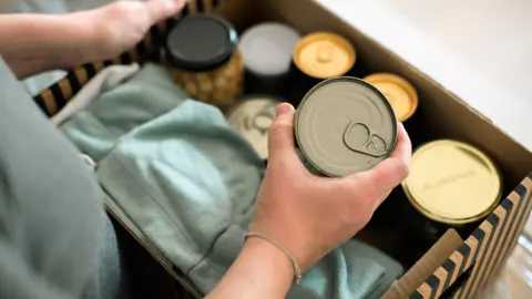 Getty Images A woman's arms are in frame. She is holding a tin of food. Beside her is a cardboard box of food in tins and glass jars.