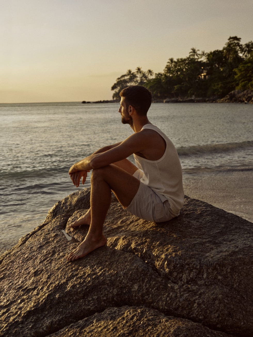 a person sitting on a rock by the beach looking out over the water during sunset