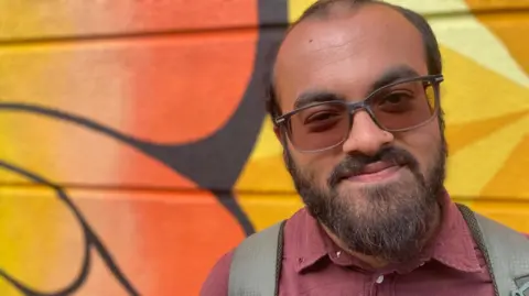 BBC / Dan Nelson A young Asian man with brown hair and a beard smiles down the camera, to the right of frame. He is wearing sunglasses, a purple shirt and a backpack. He is stood in front of a colourful wall with bright orange graffiti on it. 
