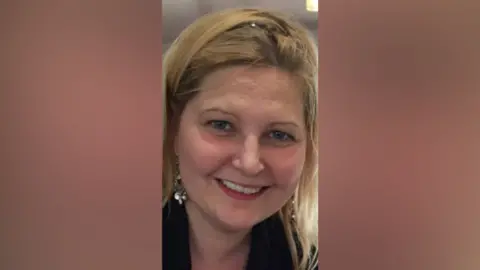 Deirdre A woman smiles into the camera. She's wearing silver earrings and wearing a black jacket. She has blond hair. 
