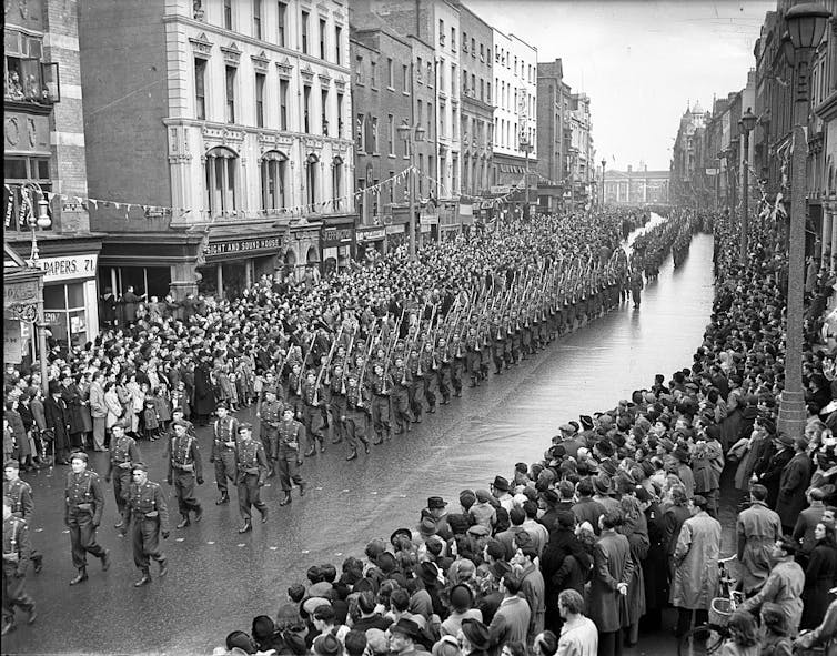 A black and white photo of people marching down a city street as crowds watch along the sides.