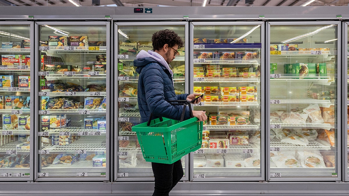 Man shopping in a supermarket while on a budget. He is looking for low prices due to inflation, standing looking at his phone in front of a row of freezers. He is living in the North East of England: new tests may be able to detect junk food consumption from blood and urine samples