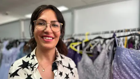BBC Stefanie Lakin stands in front of a rack of dresses covered in sequins. She is wearing a white blouse with black stars and is smiling at the camera.