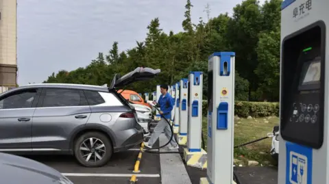 Getty Images A man charges an electric car at an electric vehicle charging station on a street in Fuyang, China. Cars are parked in front of a row of blue and white charging stations. 