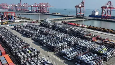Getty Images Rows and rows of stacked cars at a port in Suzhou, in eastern China's Jiangsu province, on April 7, 2025. The loading docks and cranes are visible, and beyond them the ocean.  