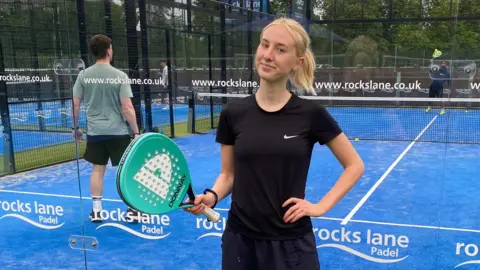Rosie Hepworth Rosie is standing in front of a blue astro padel court. She is wearing a black t-shite and matching tracksuit bottoms and we can only see her from the waist up. She is holding a turquoise padel racket in one hand and the other hand is on her hip. Rosie has shot blonde hair, which has been tied back into a low ponytail. She is smiling into the camera with no teeth. Behind her are two men playing on the court, who can be seen through the glass walls. There is white branding written on the glass.