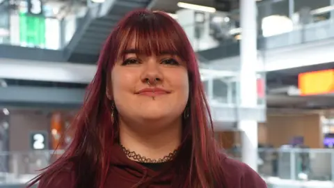 BBC A woman with long brown hair, wearing a maroon shirt and a necklace, smiles at the camera