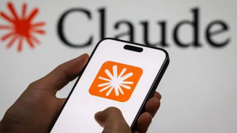 Getty Images Close up of hands holding a mobile phone displaying an orange and white icon. On the wall behind it says Claude