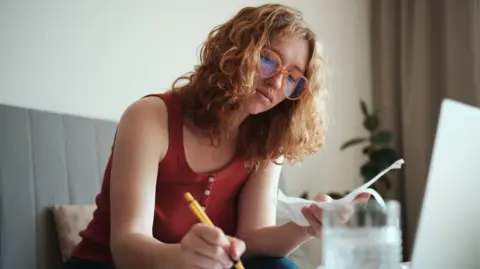 Getty Images Young woman with curly hair and glasses sits at her laptop, making notes while looking at bills