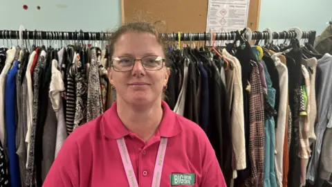 BBC Jane Platt in a bright pink polo shirt. It says Big Venture Centre. Her lanyard says the same. She has glasses and is smiling. There are lots of clothes on a big rack behind her.