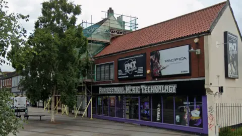 Google Maps The outside of the Professional Music Technology store in Norwich. It has a large sign with the stores title, and purple borders around the window. There is a tree to the left of it.