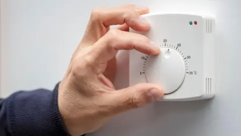 Getty Images A mans hand adjusts a white thermostat on a white wall. The mans arm is covered by a navy sweatshirt.