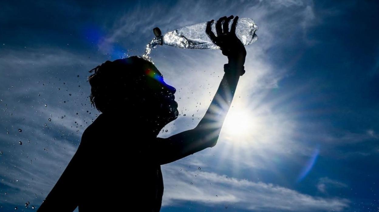A photo of a silhouette of a child pouring water over his head during a heatwave, against a background of blue sky and sunshine