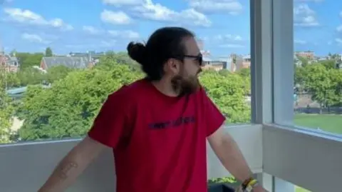 James Wickham Dylan Barber is a man, looking to the right of the image. He is wearing sunglasses, and has a beard. His hair is tied up into a bun. He is wearing a bright red tshirt, and behind him there is a white ledge, and green trees behind it. 