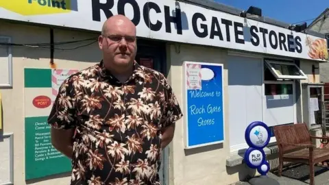 Tim Brentnall, with glasses and shaven head, stands with his hands behind his back wearing a brown, black and white Hawaiian-style shirt. In the background is his former store and Post Office, with a sign saying "Roch Gate Stores". A post office logo is visible. 