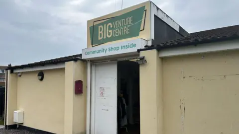 The Big Venture Centre is a single-storey yellow building. Some of the paint has chipped away. One of its white doors also looks like a little old and tired. The sign says: Big Venture Centre. Community Shop Inside.
