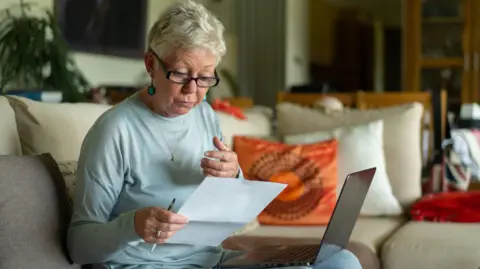 Getty Images An elderly lady looks at a bill while sitting with a laptop on her knee on a sofa