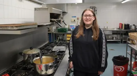 North Tyneside Council Rachel Fairless is standing smiling in the kitchen beside a big pot on a large hob. She is wearing a black top, glasses and has long fair hair.