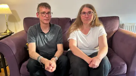 BBC Chris, a man with glasses and wearing a grey polo shirt, is sat on a purple sofa next to his wife Diana, who had long light brown hair and is wearing glasses and a white T-shirt. They are both looking at the camera