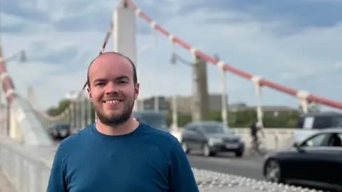 Lewis Eager Young man in blue t-shirt standing in front of a suspension road bridge with cars in the background