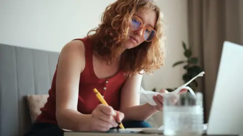 Getty Images Woman wearing glasses sits at a table with a pen in one hand and receipts in the other.