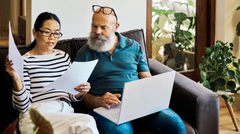 Getty Images An older couple sits together on the sofa. The woman on the left is looking at some paperwork while the man on the right has an open laptop resting on his knees. 