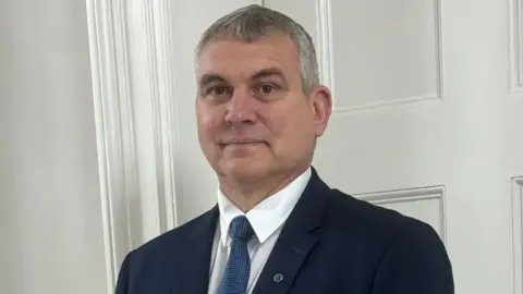 Steven Smith A headshot of a man with grey hair wearing a navy suit, white shirt and blue tie smiling into the camera. He is standing in front of a white wall. He has a small badge on his lapel.
