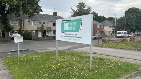 A sign for the Big Venture Centre. There is a grassy area behind with daffodils. We can also see a residential road with some houses in the background. 