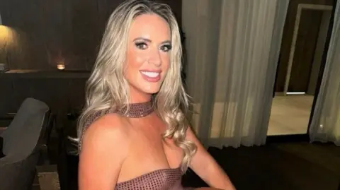 BBC A blonde woman in a dress smiles at the camera in front of chairs, tables and curtains in a dimly lit room.