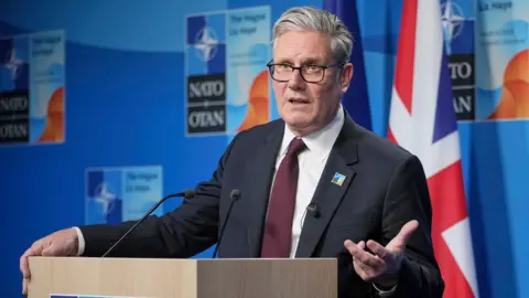 PA Media Keir Starmer gestures with his hand while speaking at a Nato summit on Wednesday in the Hague, he's wearing a suit and tie and glasses and stands at a podium with the UK flag in the background against a blue wall with the Nato logo on.