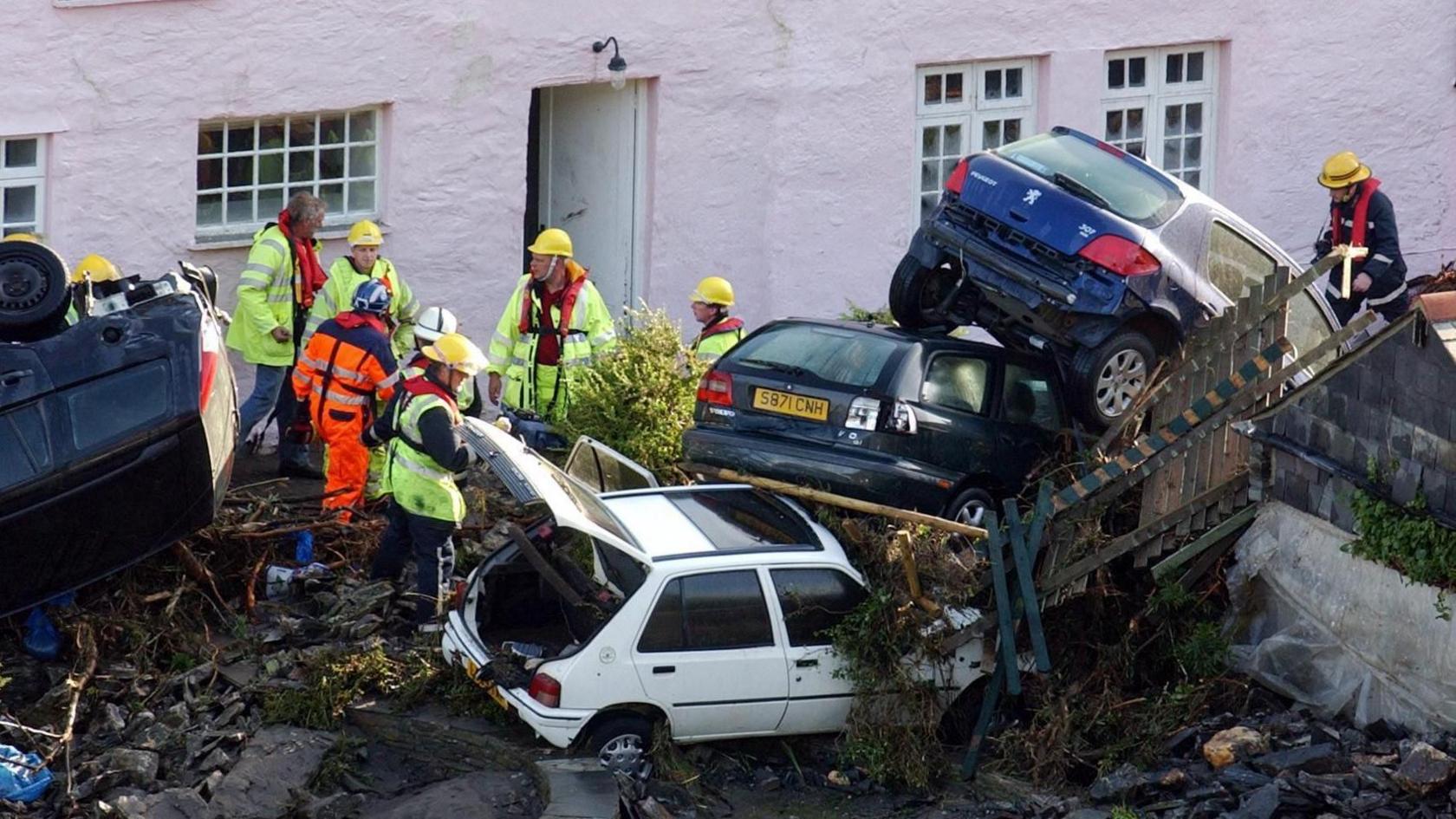 Photo of three cars piled on top of each other and emergency services personnel in front of a pink house following damage left behind following floods