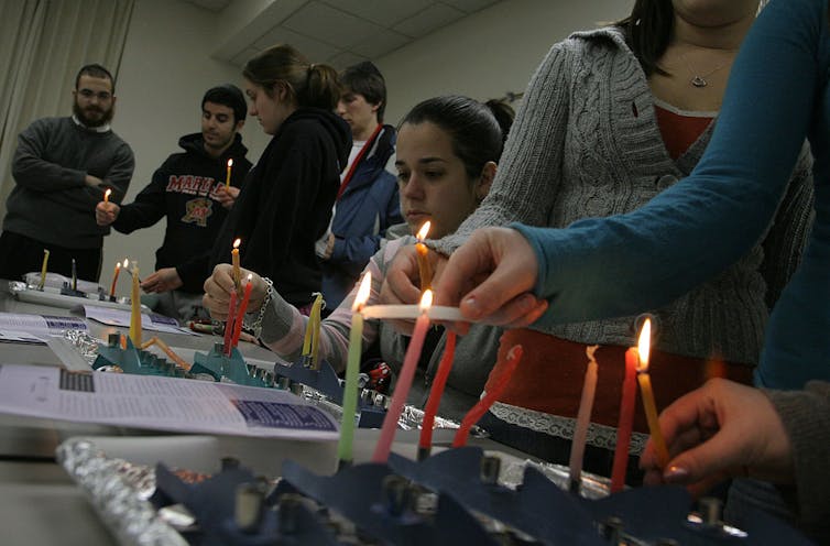 Half a dozen students sit and stand around a table where they are lighting thin colored candles.