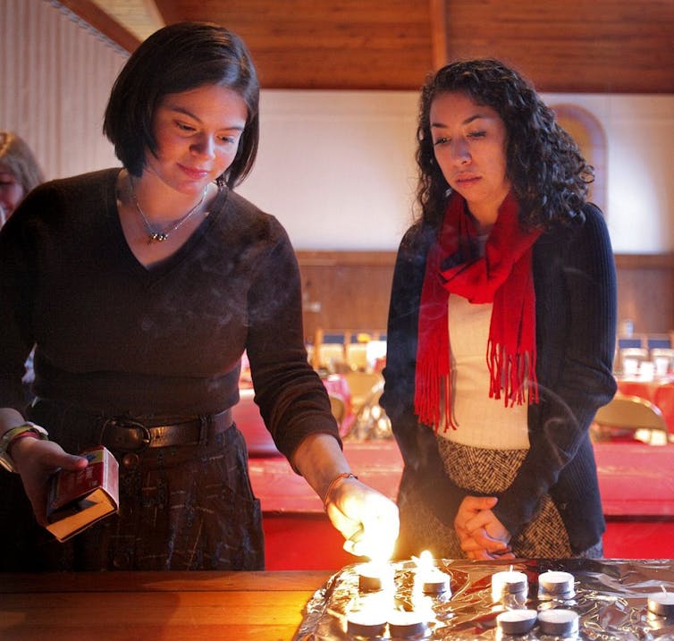 Two women in sweaters stand by a table as they light small tea candles.