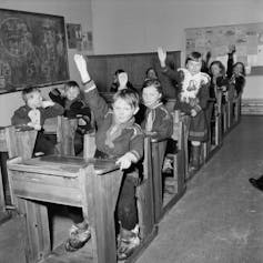 A black-and-white photo? shows about a dozen children in heavy clothing sitting at wooden desks inside.