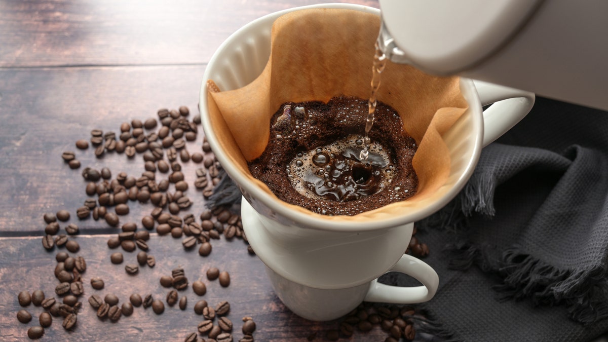 Hot water is poured into a coffee filter in a mug, beans seen scattered on table.