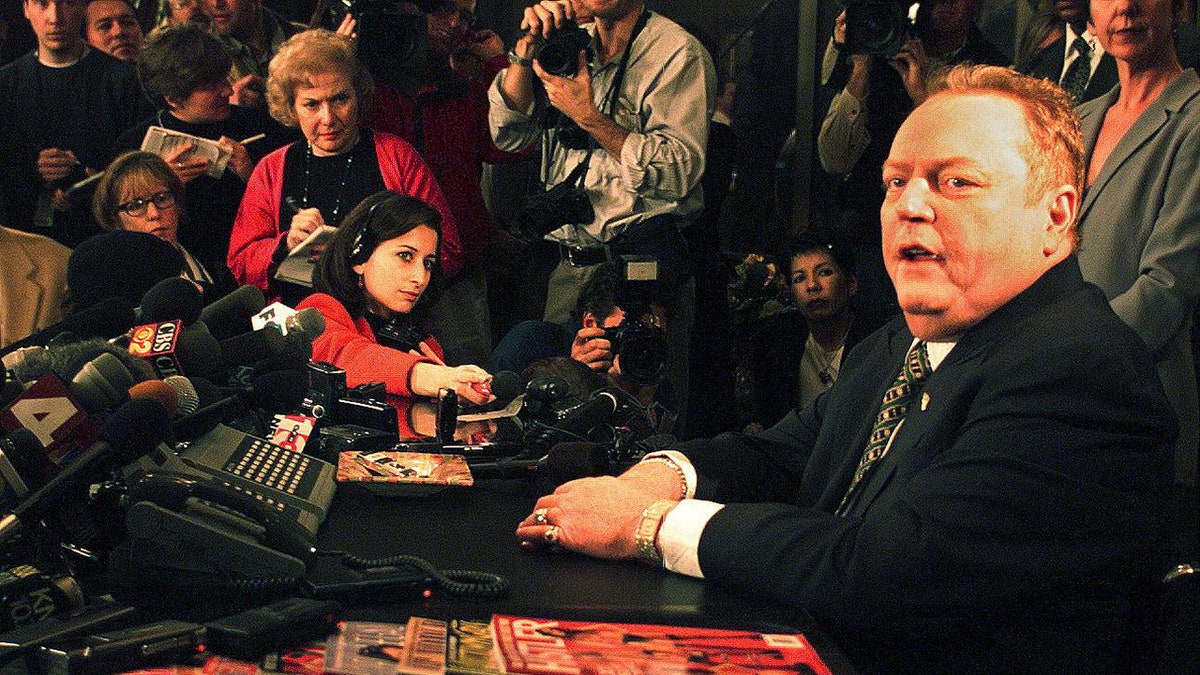 Larry Flynt looking at the camera during a press conference as the pres surrounds him.