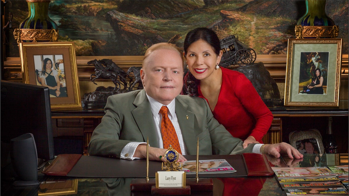 Liz Flynt wearing a red blouse leaning next to her husband Larry Flynt in a grey suit smiling sitting at his desk.