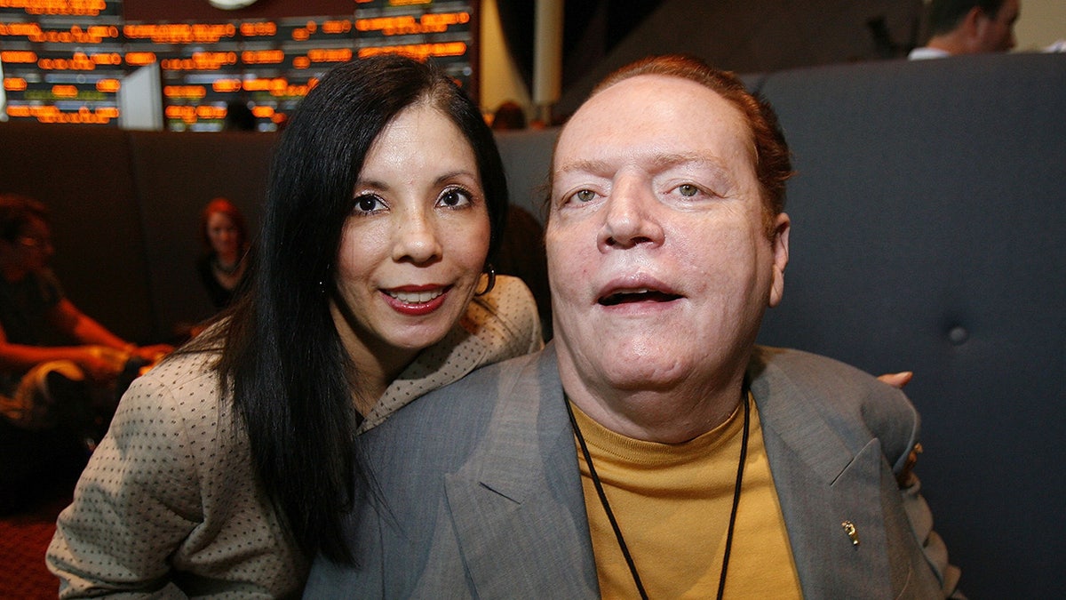 Liz Flynt leaning behind her husband Larry Flynt, who is wearing a grey blazer and a yellow shirt.