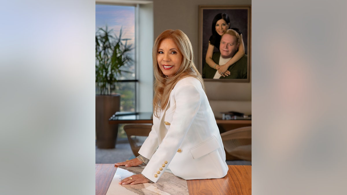 Liz Flynt wearing all white smiling in front of a portrait of her and Larry Flynt.