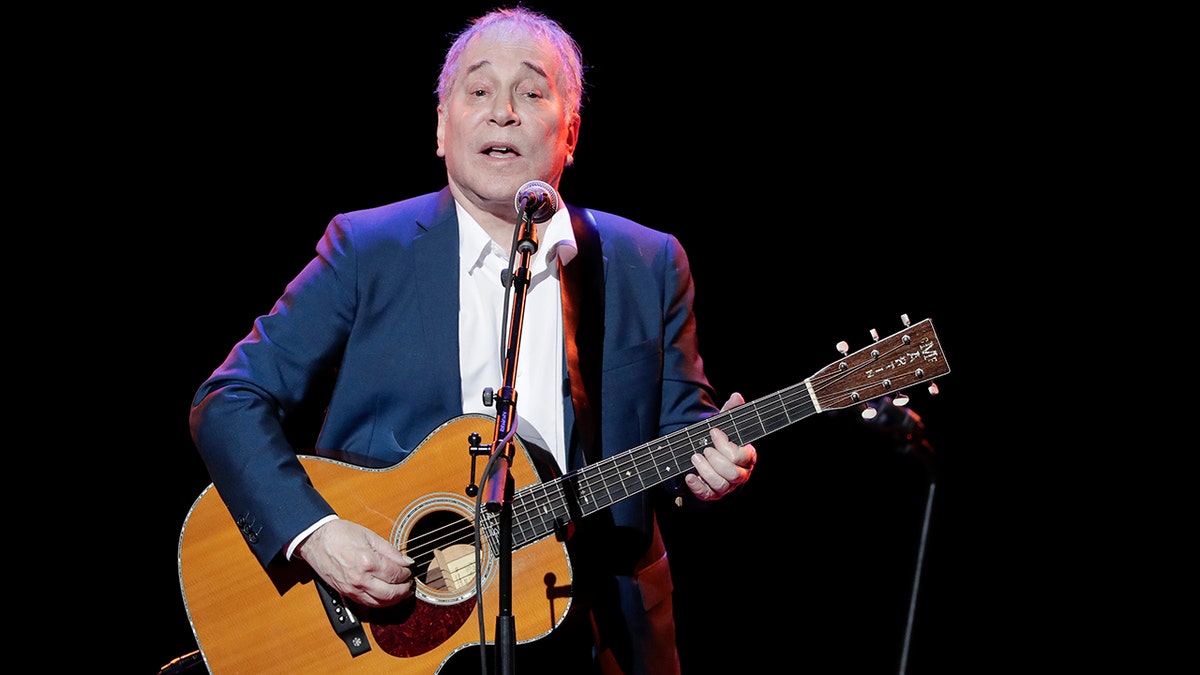 Paul Simon wears a blue blazer on stage with a guitar