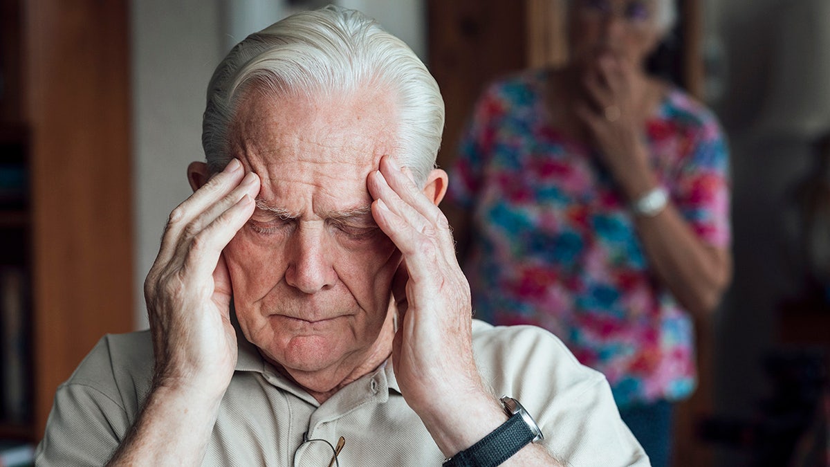 Senior man who has a chronic illness/Alzheimer's disease sitting in a living room at his home in the North East of England. He is holding his forehead with a negative expression, experiencing anxiety/pain. His wife is out of focus behind him, looking at him in worry.