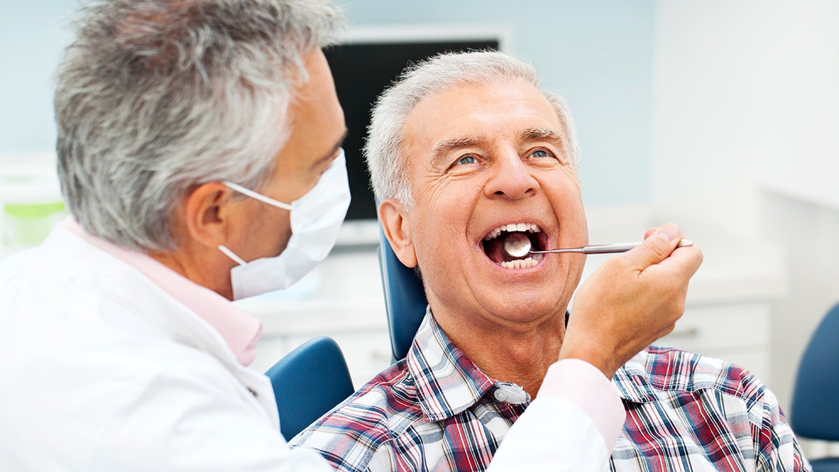 Senior man at the dentist getting his teeth examined as a dental mirror is placed into his mouth and he looks ahead sitting in his chair