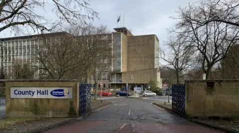 BBC An outside view of Durham County Hall from outside the gates. Cars can be seen parked outside the large grey building.