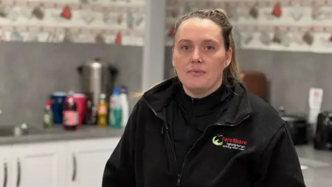 BBC Naomi Winter with brown hair tied back in a ponytail, with a nose ring, wearing a black fleece that has the words 'fareshare' on the chest, stands in a kitchen.