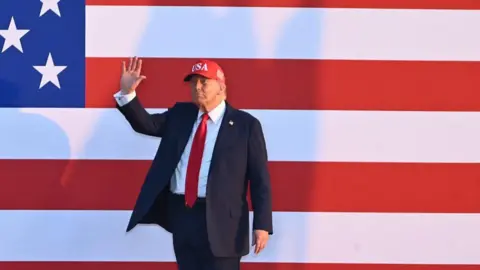 Getty Images Donald Trump waves wearing a baseball cap and suit in front of a huge US flag poster