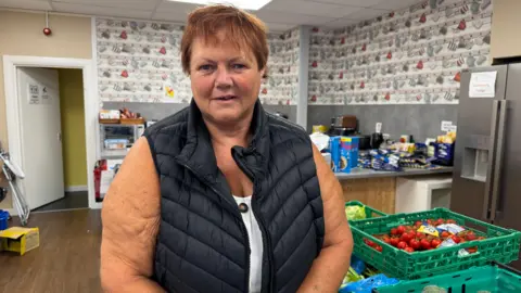 Diane Snedker, wearing a black gilet, with short red hair, stands next to boxes of food in a kitchen. 