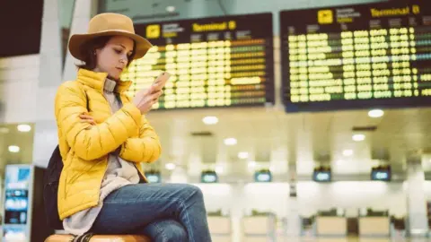 Getty Images A lady in a hat and yellow jacket sat in front of airport departure boards, sat on her suitcase and looking in to her mobile phone.