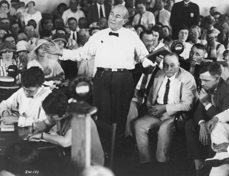A man in a white shirt and black bow tie stands with arms outstretched while talking, while others seated in rows around him look on.