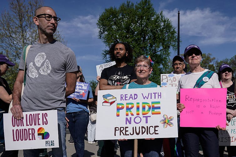 A handful of people stand outside holding placards with messages including 'Read with pride not prejudice.'